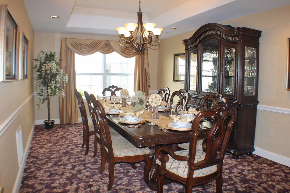 A formal dining room with a long dark wooden table set for eight people with white plates, bowls, and glasses. The room features a patterned carpet, beige walls with white trim, a large window with beige curtains, a chandelier overhead, a tall wooden china cabinet filled with glassware, and a potted plant in the corner.