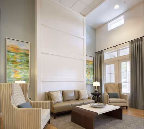 Modern senior living lounge with beige sofa, armchairs, coffee table, and large windows.