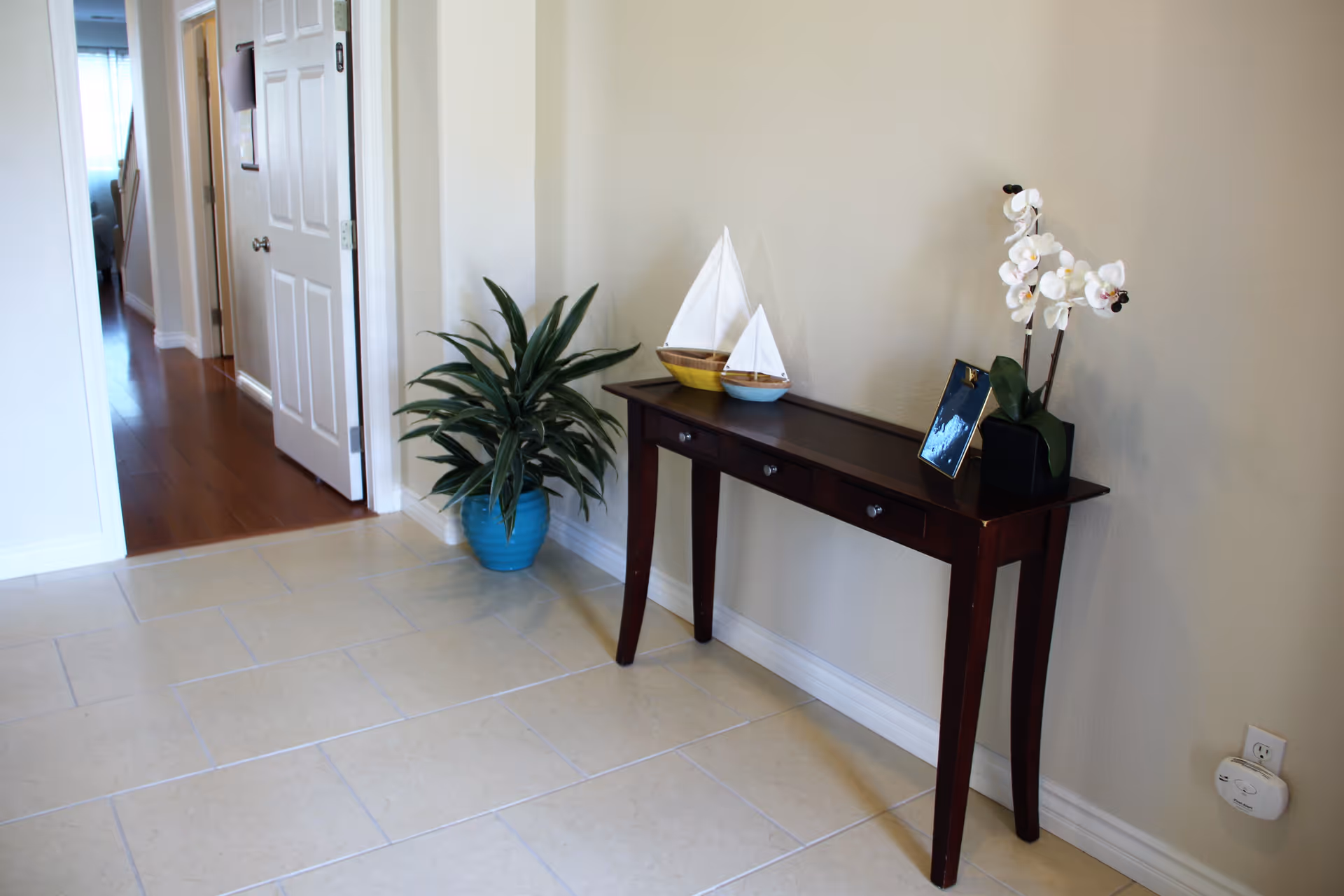 A hallway area with beige tiled floor and cream-colored walls. A dark wooden console table with four drawers is placed against the wall, decorated with two small sailboat models, a framed photo, and a white orchid plant in a black pot. Next to the table is a green leafy plant in a blue pot. An open door reveals a room with wooden flooring and a glimpse of furniture inside.