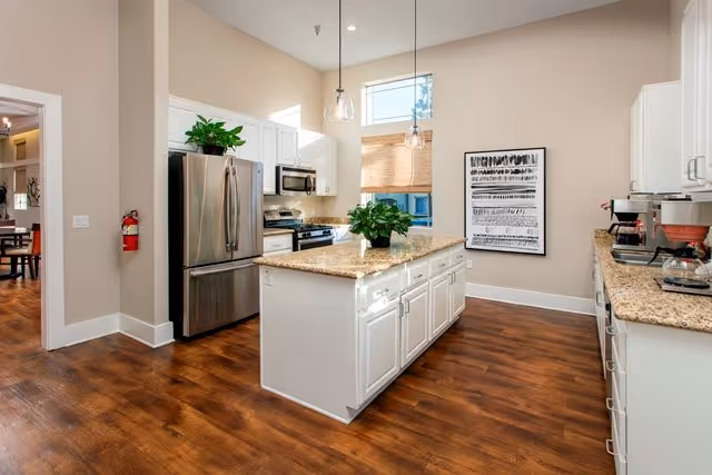 A modern kitchen with white cabinets, granite countertops, and stainless steel appliances including a refrigerator and microwave. There is a kitchen island in the center with a green plant on top. The floor is wood, and there are two pendant lights hanging from the ceiling. A window with a bamboo shade and a framed abstract artwork are visible on the far wall.