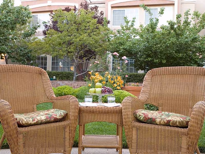 Two wicker chairs with floral cushions facing each other with a small wicker table between them holding a plate of fruit and two glasses of water, set in a garden area with green grass, flowering plants, and trees in front of a building with windows.