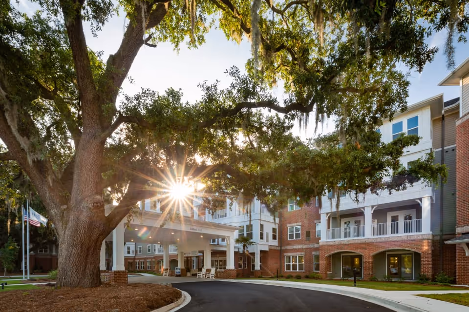 Exterior view of a senior living facility named Harmony at Savannah with a large tree in the foreground, sunlight shining through the branches, and a covered entrance with rocking chairs. The building is multi-story with brick and siding, featuring balconies and large windows.
