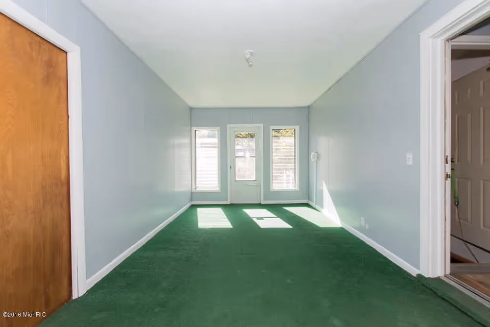 Empty narrow room with green carpet, light gray walls, a wooden door on the left, and a white door on the right leading to another room. At the far end, there are two tall windows flanking a glass door that lets in natural light.