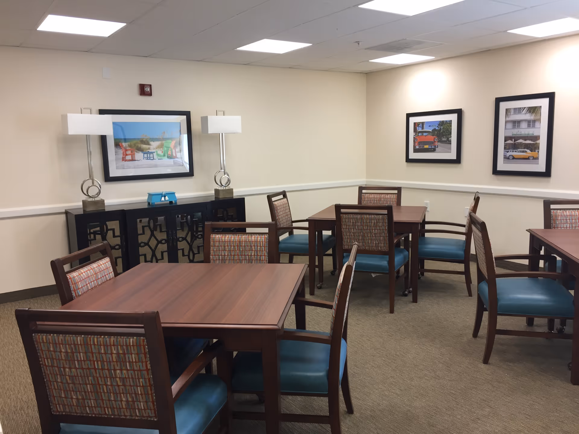A dining room with several wooden tables and chairs with blue cushions and patterned backs. The walls are beige with three framed pictures hanging, and there are two modern table lamps on a black sideboard against the wall.