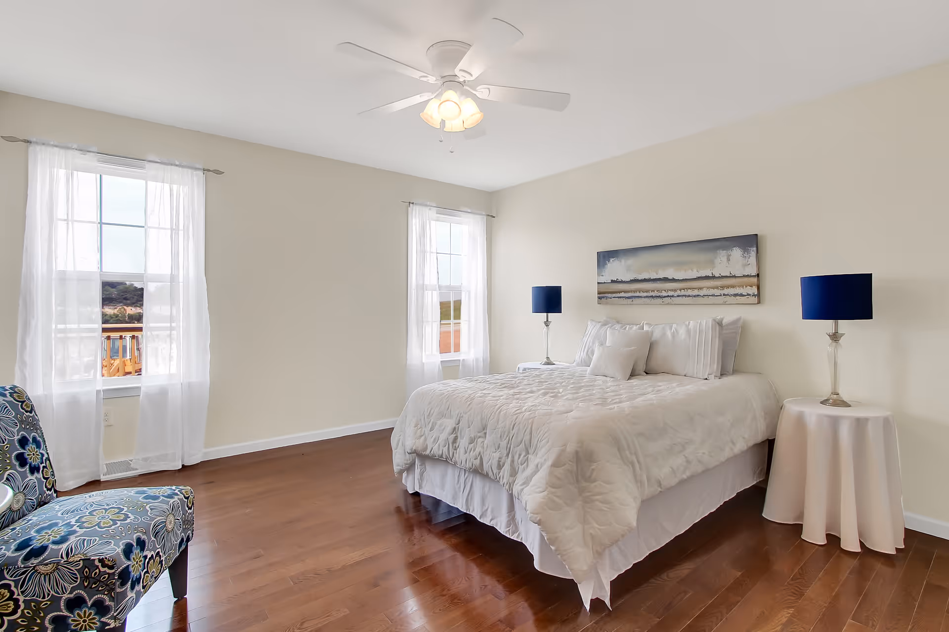 A bright bedroom with two large windows covered by sheer white curtains, a bed with white bedding and pillows, two bedside tables each with a blue lamp, a floral patterned chair, wooden floor, and a ceiling fan with lights.