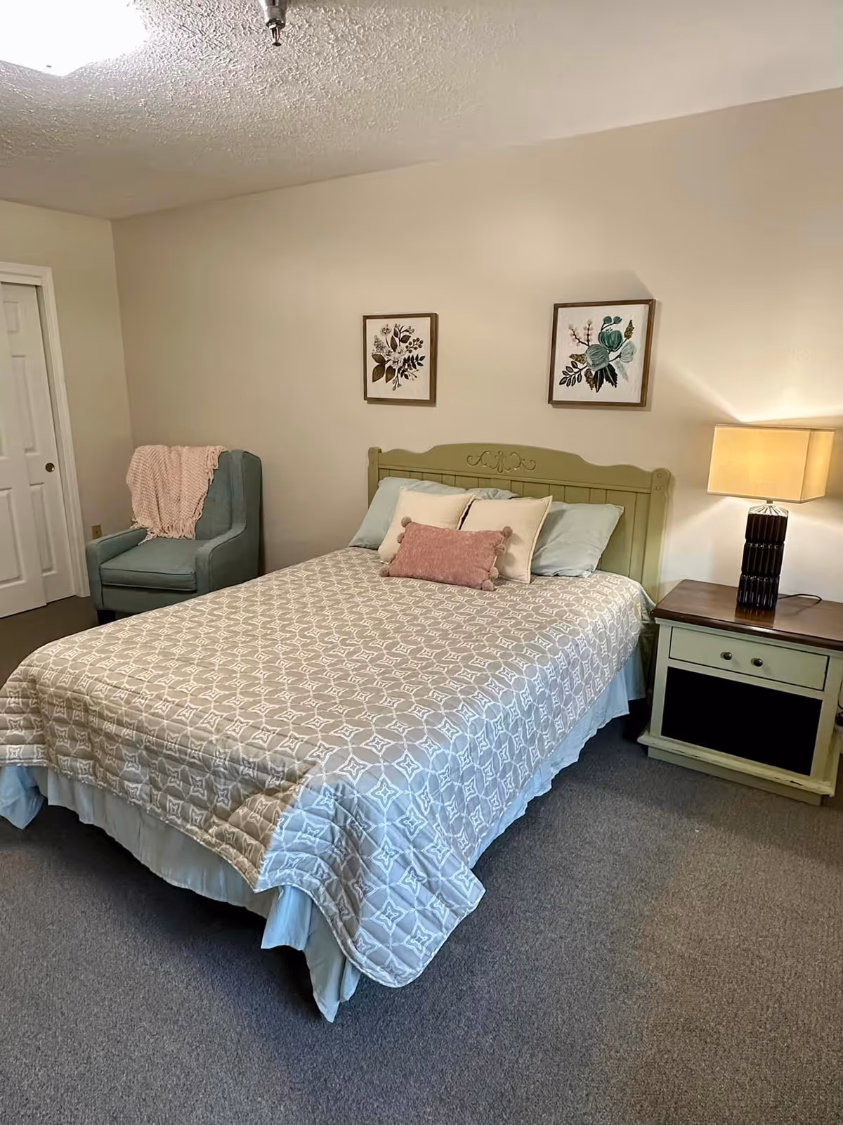 A cozy bedroom with a bed featuring a patterned quilt and multiple pillows. There is a green headboard, a nightstand with a lamp on the right side of the bed, and a blue armchair with a pink throw blanket in the corner. Two framed botanical prints hang on the wall above the bed.