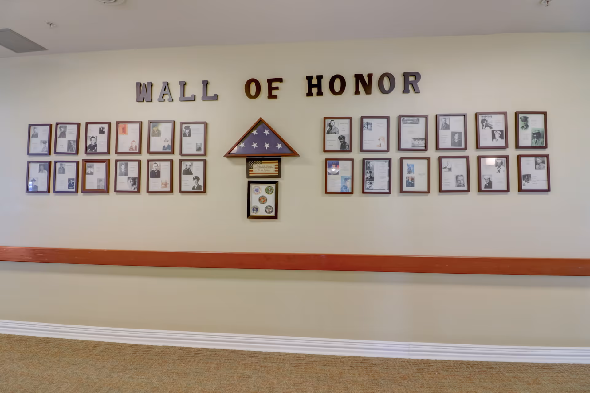 A wall display titled 'Wall of Honor' featuring framed photographs and documents honoring individuals, with a folded American flag and military insignia in the center, mounted on a beige wall above a wooden handrail.