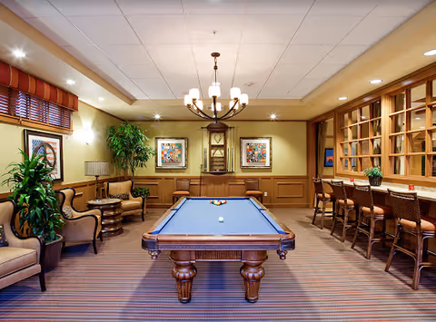 Indoor recreation room with a pool table centered beneath a chandelier, surrounded by seating, bar stools, plants, and framed artwork.