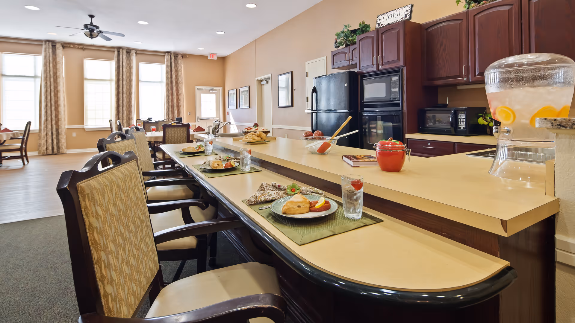 Interior view of a senior living facility dining area with a kitchen counter and bar stools. The counter has plates with food, glasses of water with strawberries, and a large beverage dispenser with lemon water. The kitchen area features dark wood cabinets, a refrigerator, microwave, and oven. In the background, there are tables and chairs near large windows with curtains.