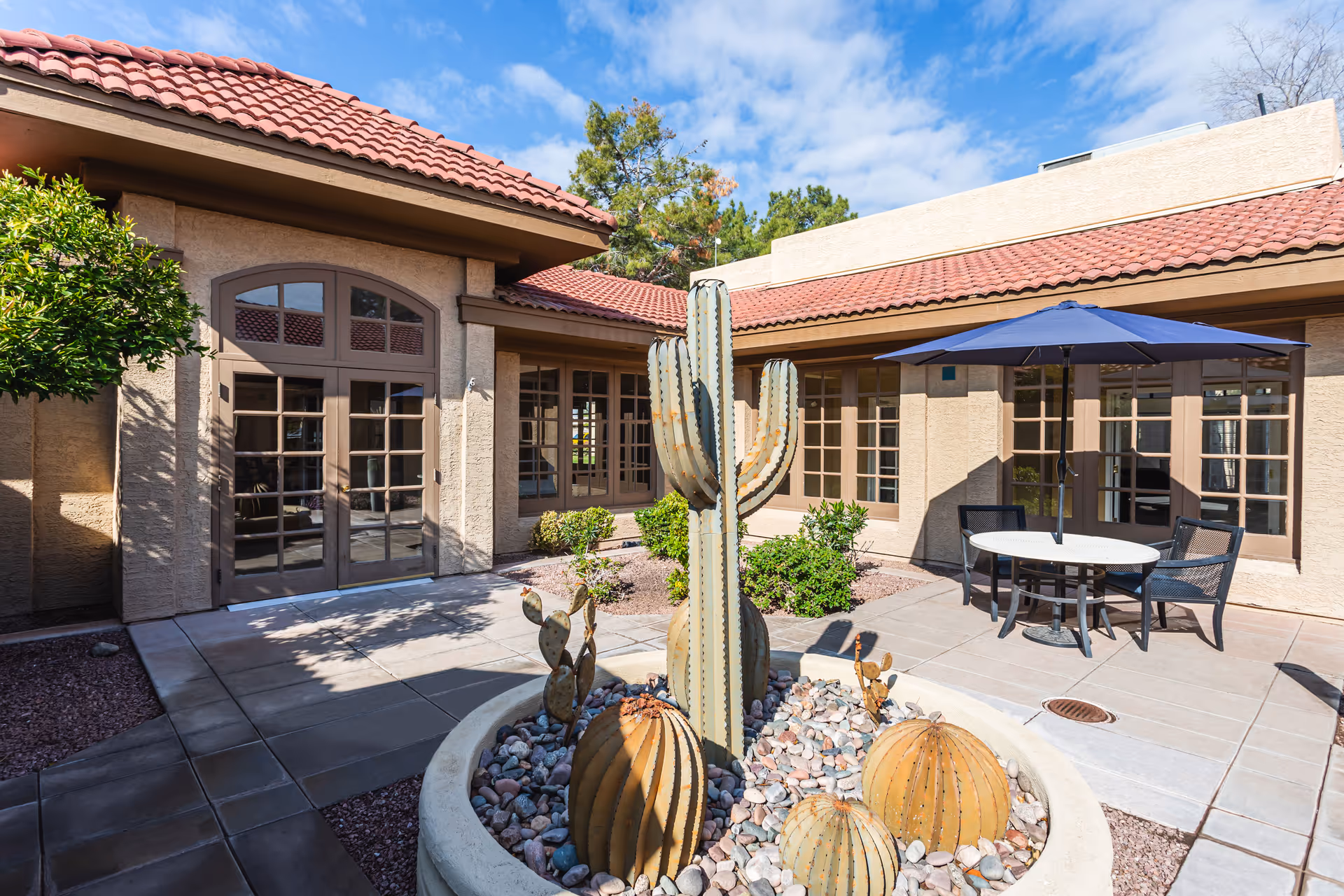 Outdoor courtyard area at a senior living facility with a large cactus centerpiece surrounded by rocks, a round table with four chairs and a blue umbrella, beige stucco walls, multiple windows, and a red tile roof under a partly cloudy sky.