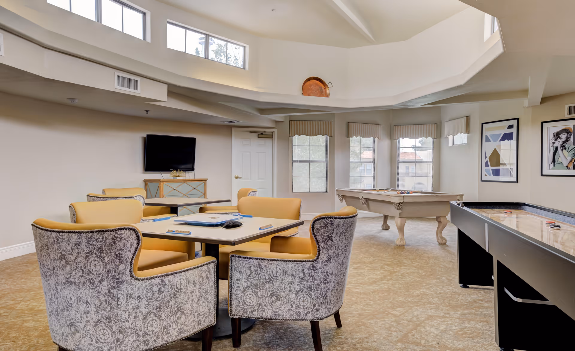 A bright and spacious game room with a table and six upholstered chairs arranged around it. The room features a pool table and an air hockey table, with large windows letting in natural light. There is a flat-screen TV mounted on the wall and framed artwork hanging on another wall. The ceiling has a unique architectural design with high windows near the top.