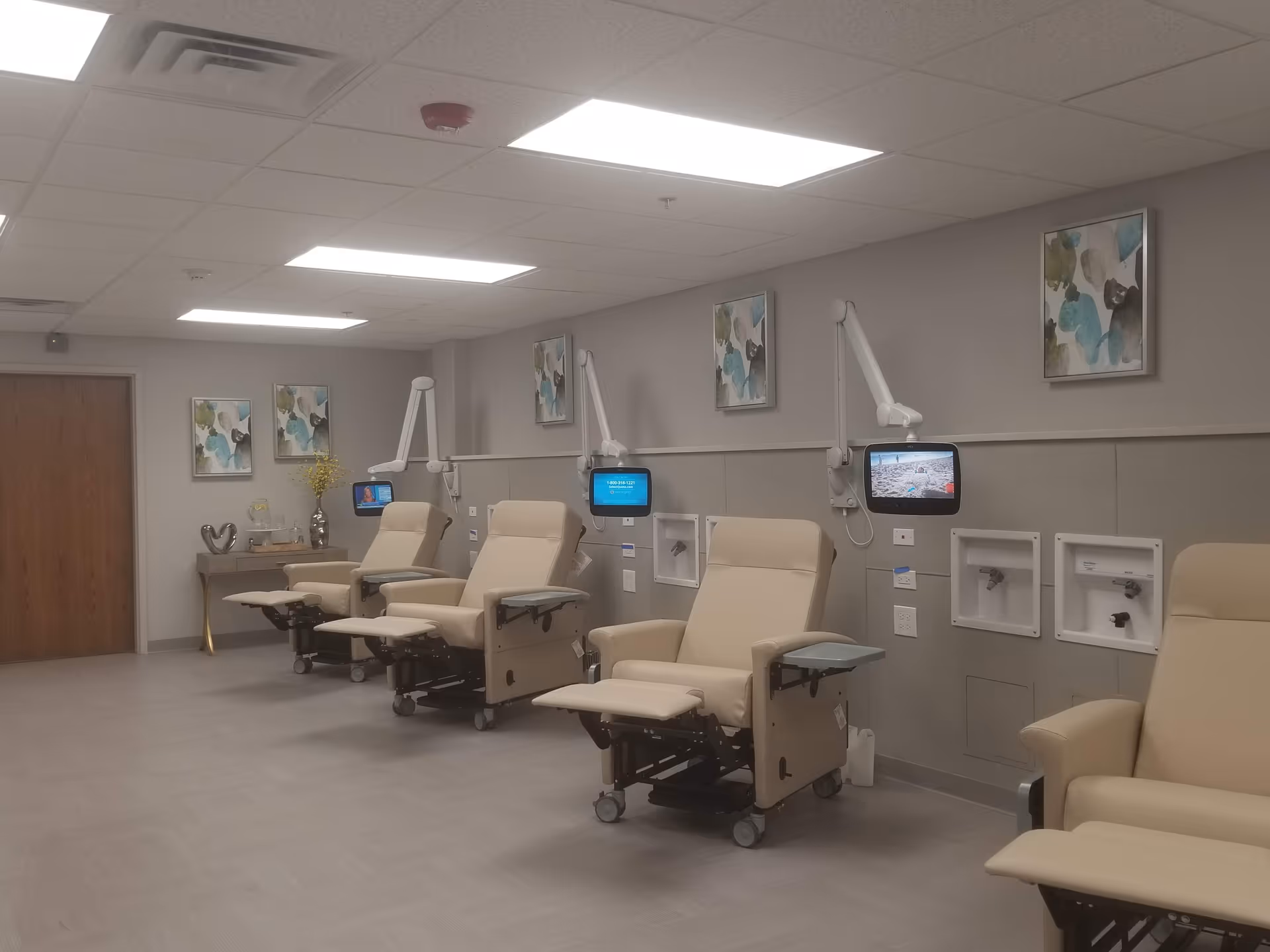 A clinical treatment room with a row of beige reclining chairs, wall-mounted monitors, and medical hookups.