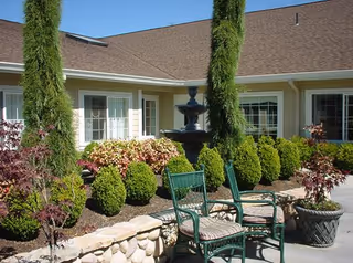Outdoor courtyard area with green metal chairs and a small black fountain surrounded by neatly trimmed bushes, potted plants, and a stone border in front of a beige building with white-framed windows under a clear blue sky.