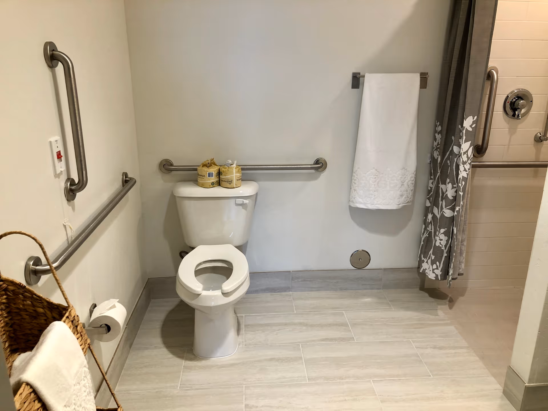 Accessible bathroom featuring a toilet with raised seat, multiple grab bars, a towel on a wall rack, and a shower with a floral curtain.