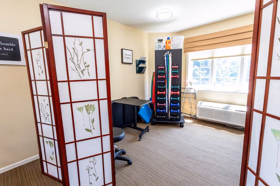 A bright room with a large window covered by a beige blind, a white air conditioning unit below the window, and a walker nearby. In the center, there is a rack holding colorful dumbbells and resistance bands. A black desk and a black rolling stool are positioned against the wall. The room is partially divided by a decorative folding screen with floral designs. The walls have motivational signs, including one that says 'choose happy.'