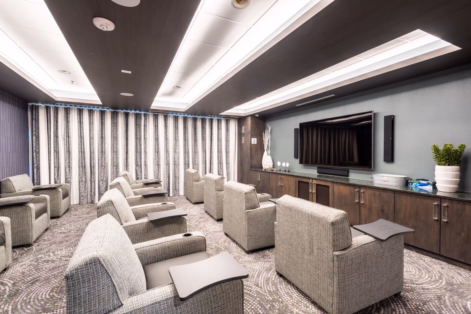 A modern media room with multiple comfortable armchairs arranged in rows facing a large flat-screen TV mounted on a wall with built-in cabinets below. The room features a patterned carpet, a ceiling with recessed lighting, and curtains covering the windows.