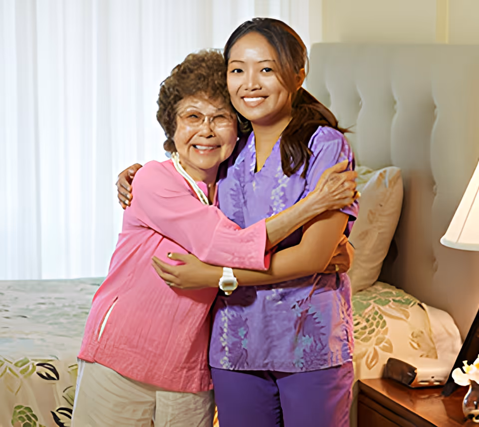 An elderly woman in a pink top and beige pants hugging a younger woman in purple scrubs in a bedroom with a bed, pillows, and a bedside lamp.