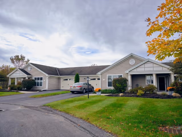 Exterior view of a single-story residential building with beige siding, white trim, and a dark roof. There is a driveway with a parked car, well-maintained green lawns, shrubs, and a tree with autumn-colored leaves on the right side. The sky is partly cloudy.