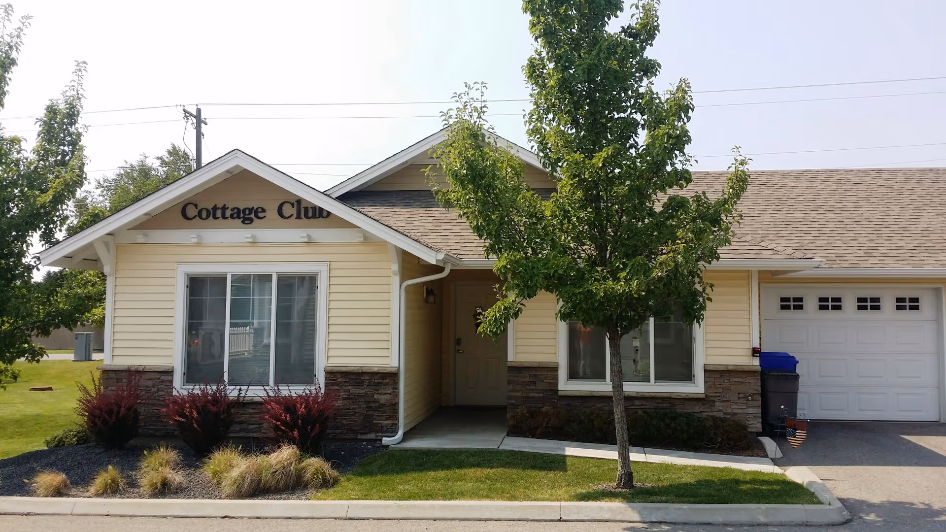 Exterior view of a single-story cottage building with beige siding and stone accents. The building has a front entrance with a small covered porch, two windows, and an attached garage. There is a tree and landscaped bushes in front of the building. The sign above the window reads 'Cottage Club'.