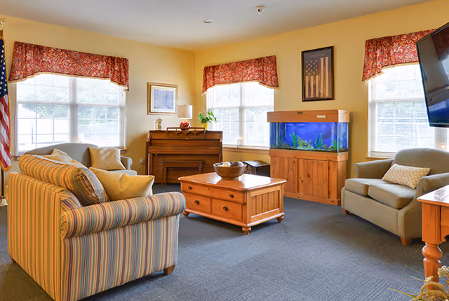 A cozy living room with a striped sofa, an armchair, a wooden coffee table, a piano, and a large fish tank. The room has three windows with red patterned valances, a framed picture on the wall, and a television mounted on the right side.