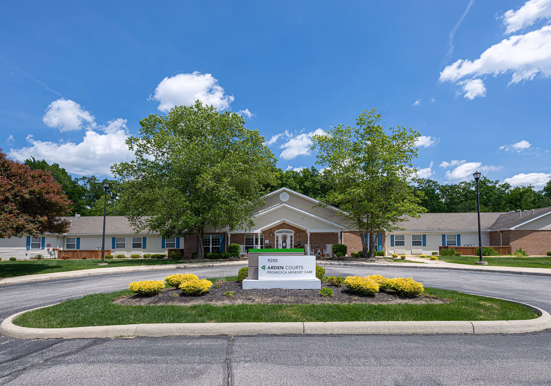 Front view of Arden Courts ProMedica Memory Care, a single-story building with a circular driveway, landscaped island and entrance sign under a blue sky.