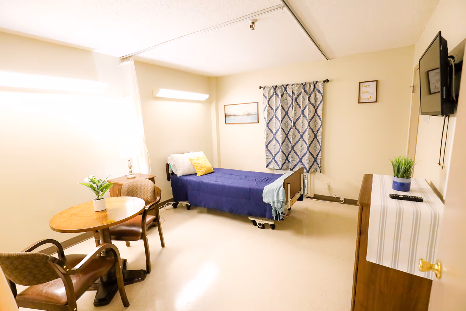 A simple, clean bedroom in a healthcare facility with a single bed covered in a blue blanket and a yellow pillow. There is a small round wooden table with two chairs, a potted plant on the table, a dresser with a striped cloth and a small plant, and a wall-mounted TV. The room has beige walls, a patterned curtain, and framed artwork on the walls.