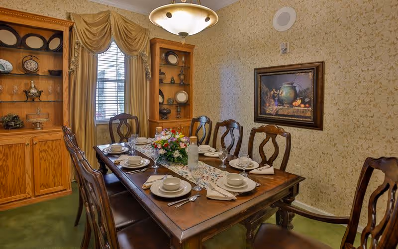 A formal dining room with a wooden table set for six people, including plates, bowls, glasses, and napkins. The table has a floral centerpiece and a decorative runner. The room features two wooden display cabinets with decorative items, a window with gold curtains, patterned wallpaper, a framed painting on the wall, and a ceiling light fixture.