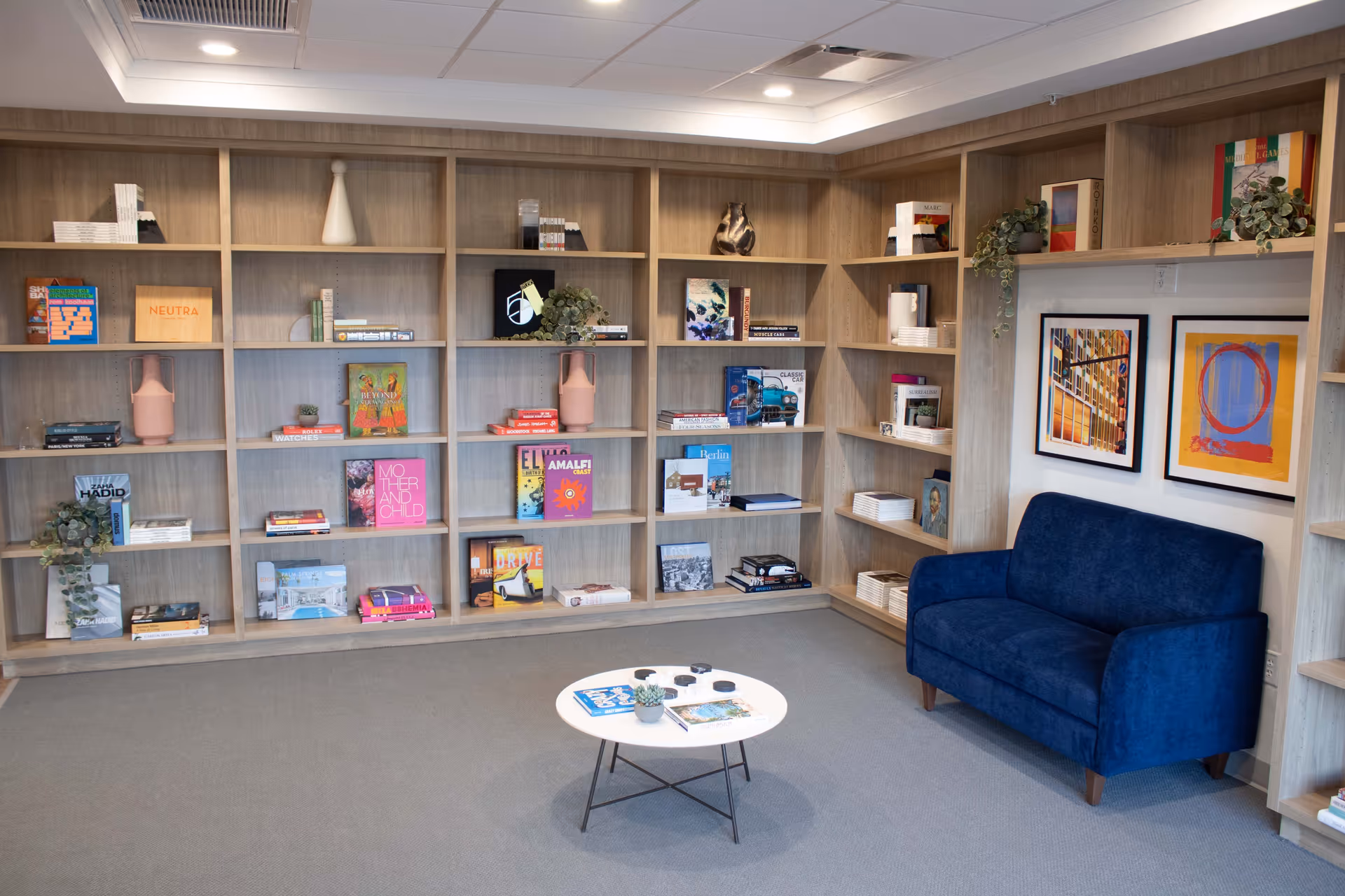 A cozy reading or lounge area with light wood bookshelves filled with books, decorative items, and plants. A small round white table with books and a small plant sits on a gray carpeted floor. A blue upholstered loveseat is positioned against the wall, which is decorated with two colorful framed artworks.