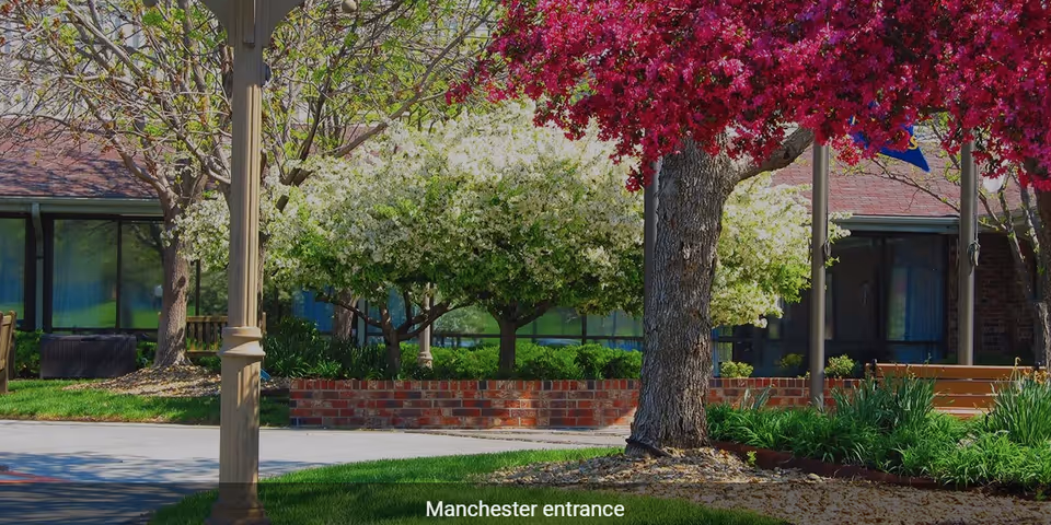 Outdoor view of Aldersgate Village Life Plan Community entrance with blooming trees, green shrubs, a brick planter, and a building with large windows in the background.