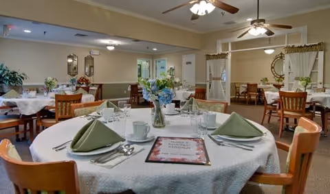 A dining room with round tables covered in white tablecloths, each set with green folded napkins, plates, cups, and silverware. There are wooden chairs around the tables, floral centerpieces, and ceiling fans with lights. The room has beige walls with decorative mirrors and curtains.