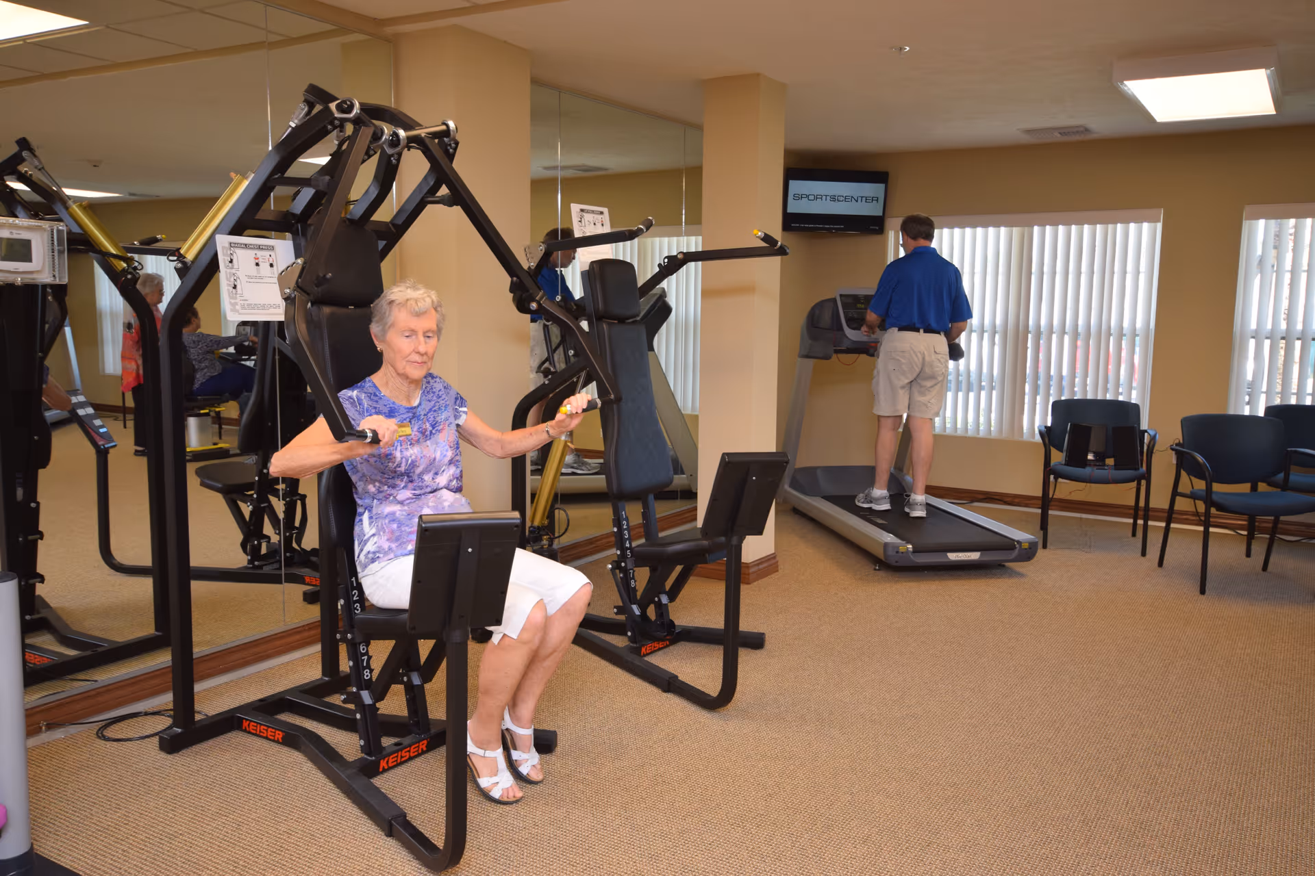 An elderly woman using a strength training machine in a fitness room while an elderly man walks on a treadmill in the background. The room has beige walls, large windows with vertical blinds, and several chairs along the wall.