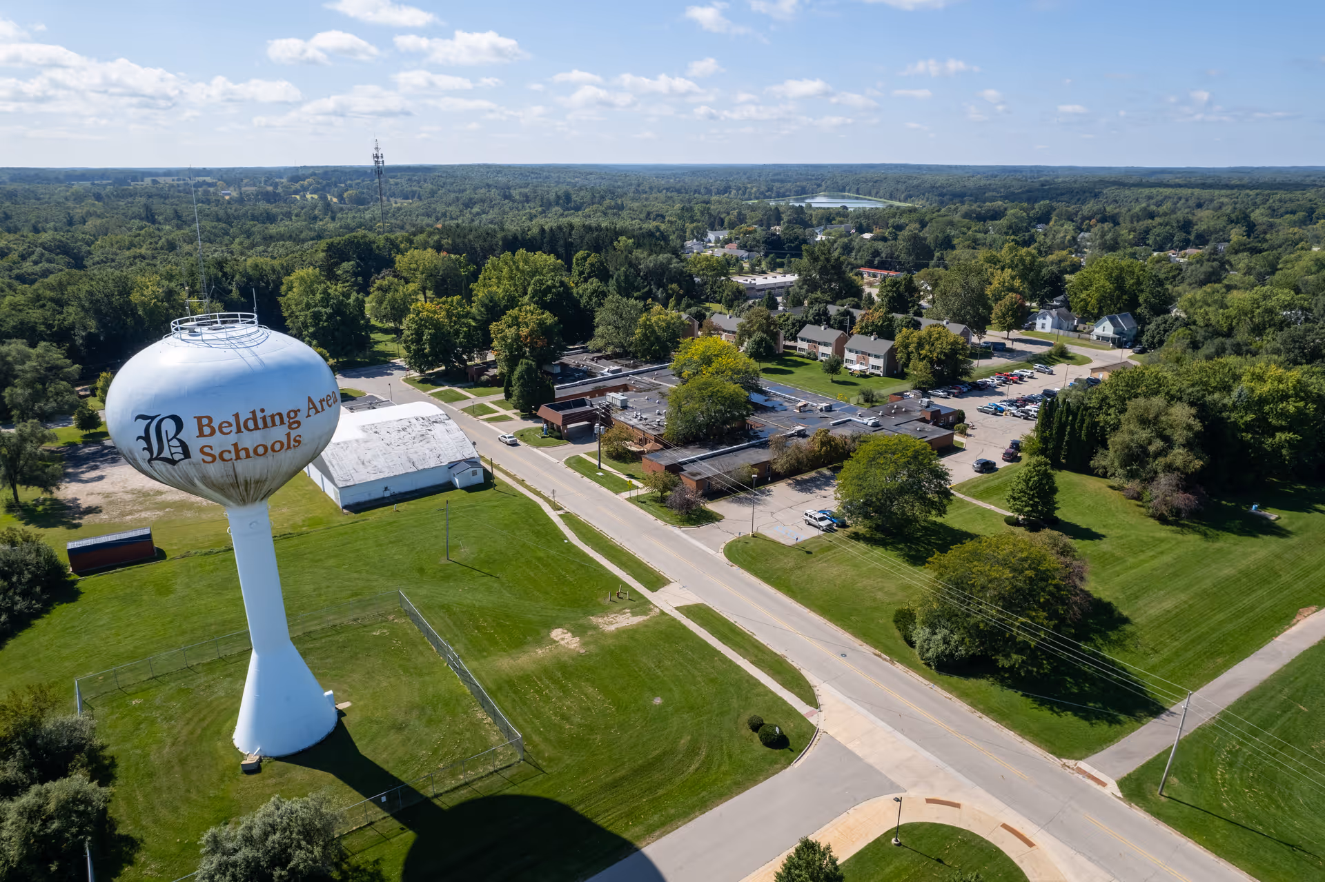 Aerial view of a suburban area with a large white water tower labeled 'Belding Area Schools' on a grassy field. Surrounding the water tower are roads, buildings, parked cars, and many trees extending into the distance under a partly cloudy sky.