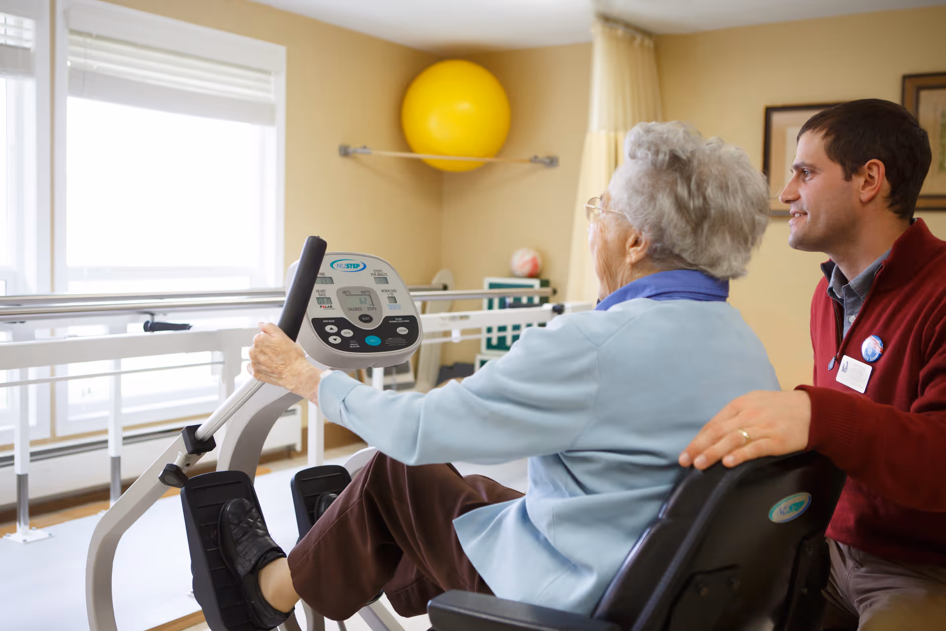 An elderly woman using a seated exercise machine with the assistance of a male caregiver in a well-lit room with large windows and exercise equipment in the background.