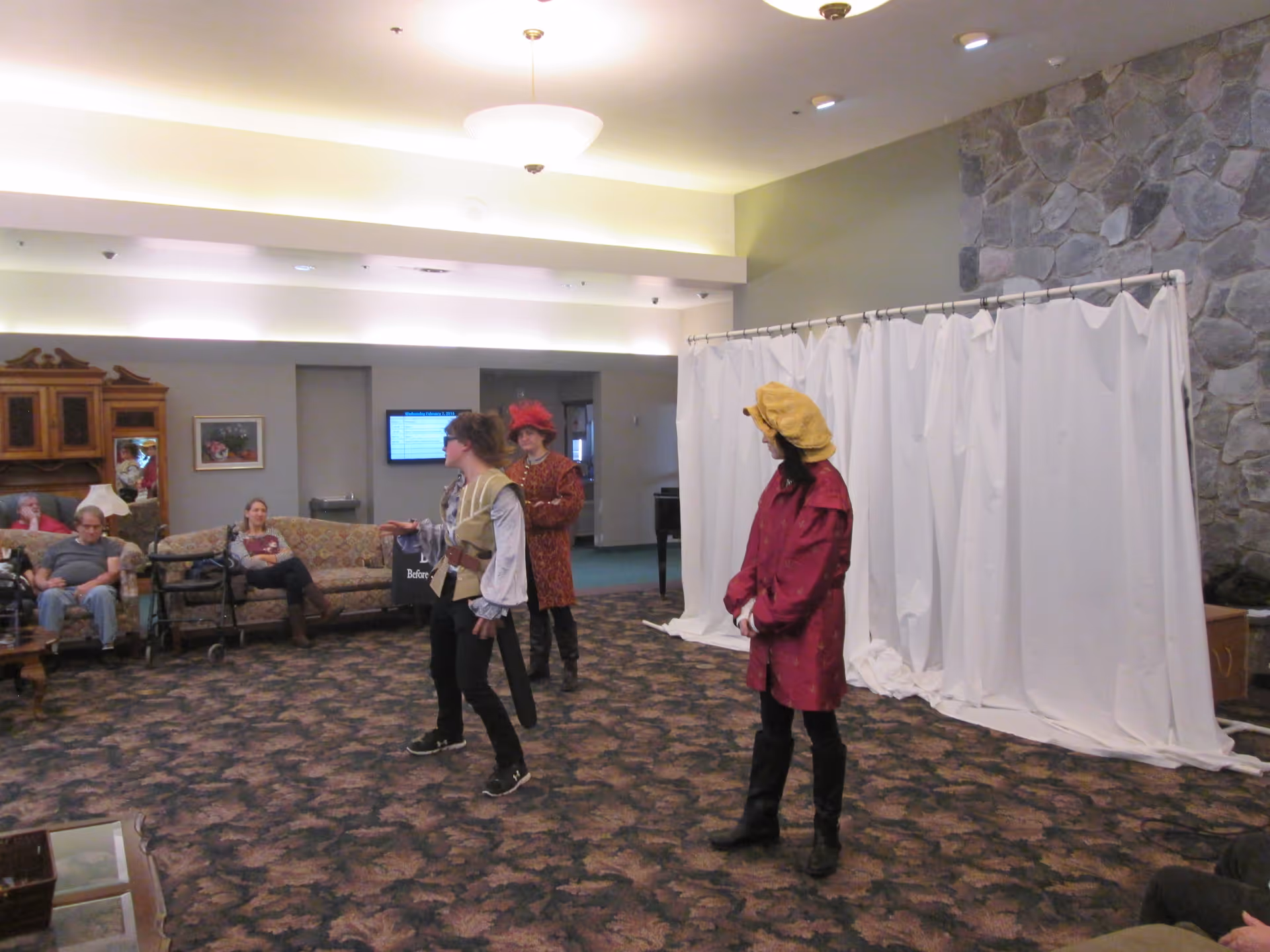 A group of people in a common area of an assisted living facility. Three individuals are dressed in historical costumes performing or interacting, while several elderly residents sit on couches and chairs watching. The room has patterned carpet, a stone wall, and a white curtain backdrop.