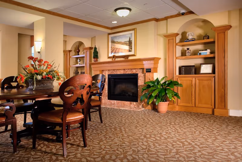 A cozy interior room featuring a round wooden table with ornate chairs, a floral centerpiece, a fireplace with a marble surround, built-in wooden shelves with decorative items, a potted plant on the carpeted floor, and a framed landscape painting above the fireplace.