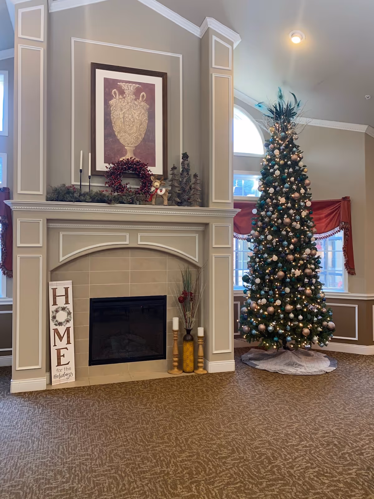 A decorated indoor living area featuring a fireplace with a framed artwork above it, holiday decorations including a wreath, candles, and a sign that reads 'HOME for the Holidays'. To the right of the fireplace is a tall Christmas tree adorned with lights and ornaments. The room has beige walls with white trim and carpeted flooring.