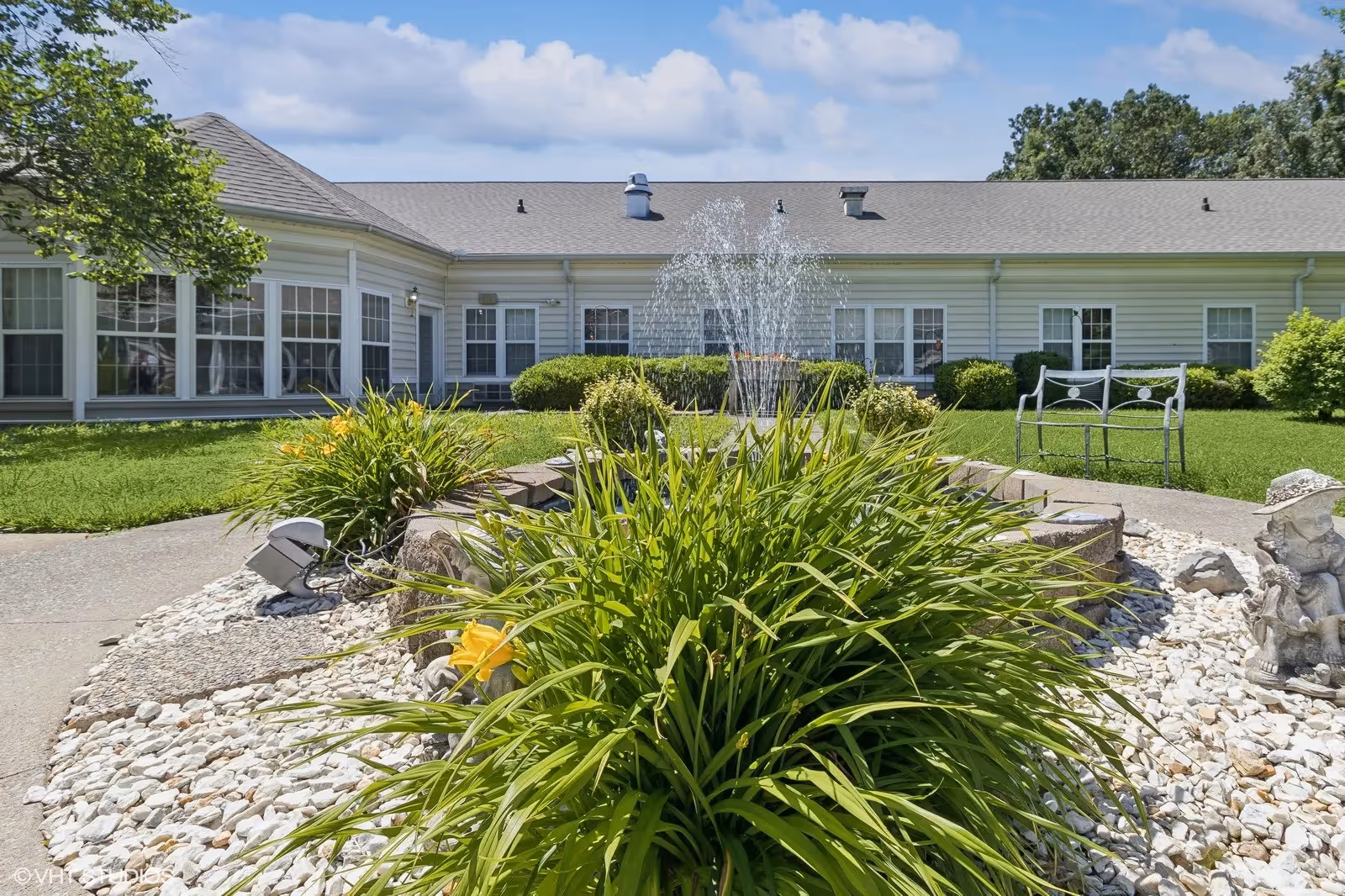 Courtyard with a central fountain, flowering plants and benches in front of a single-story building under a blue sky.