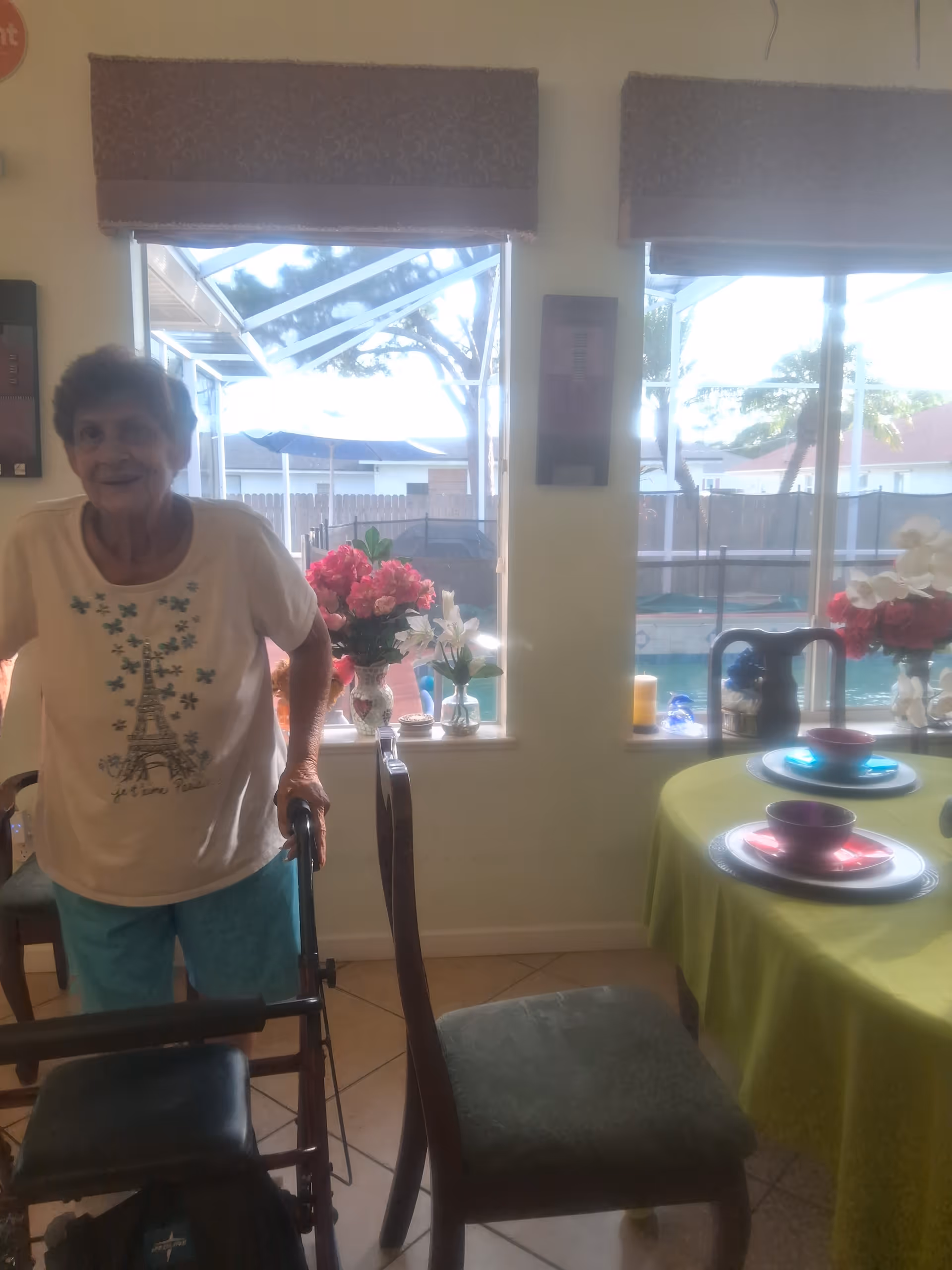 A woman using a walker stands in a dining area by windows with a table set with colorful dishes and floral arrangements and a pool visible outside.