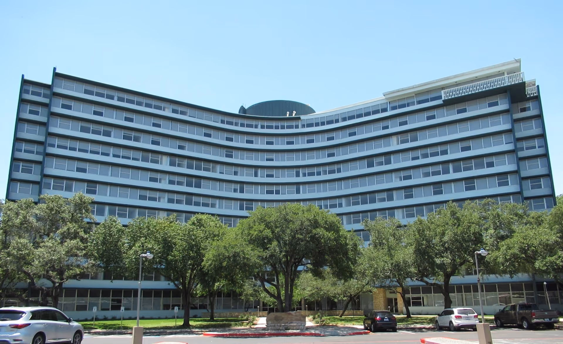 Curved multi-story building with rows of windows, trees in front and cars parked along the street.