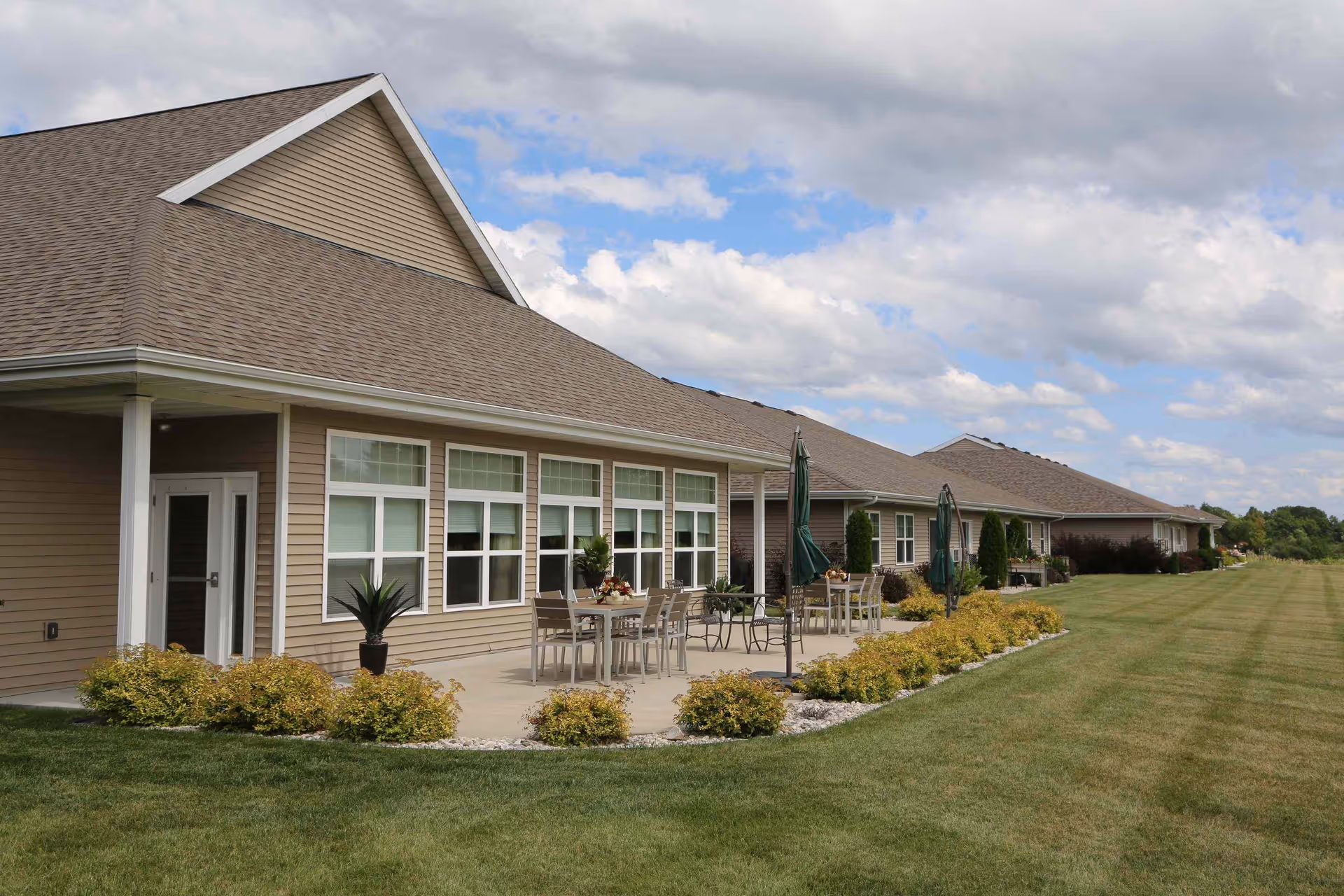 Exterior view of a single-story senior living facility building with beige siding and a brown shingled roof. There is a concrete patio area with tables, chairs, and closed green umbrellas. The patio is bordered by small bushes and landscaping rocks. The building is surrounded by a large, well-maintained grassy lawn under a partly cloudy sky.