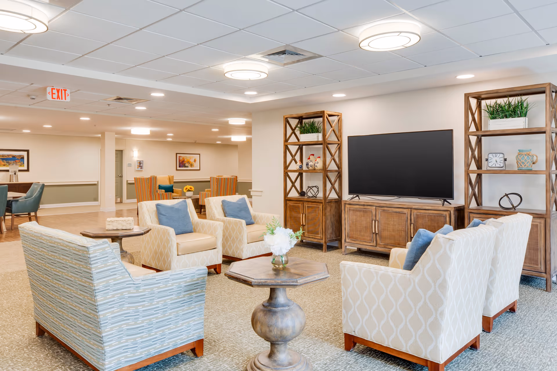 A bright and spacious senior living facility common area with comfortable armchairs arranged around a wooden coffee table. The room features a large flat-screen TV on a wooden cabinet flanked by two wooden shelving units with decorative items and plants. The ceiling has recessed lighting and circular light fixtures. In the background, there are additional seating areas with chairs and tables, artwork on the walls, and an exit sign.