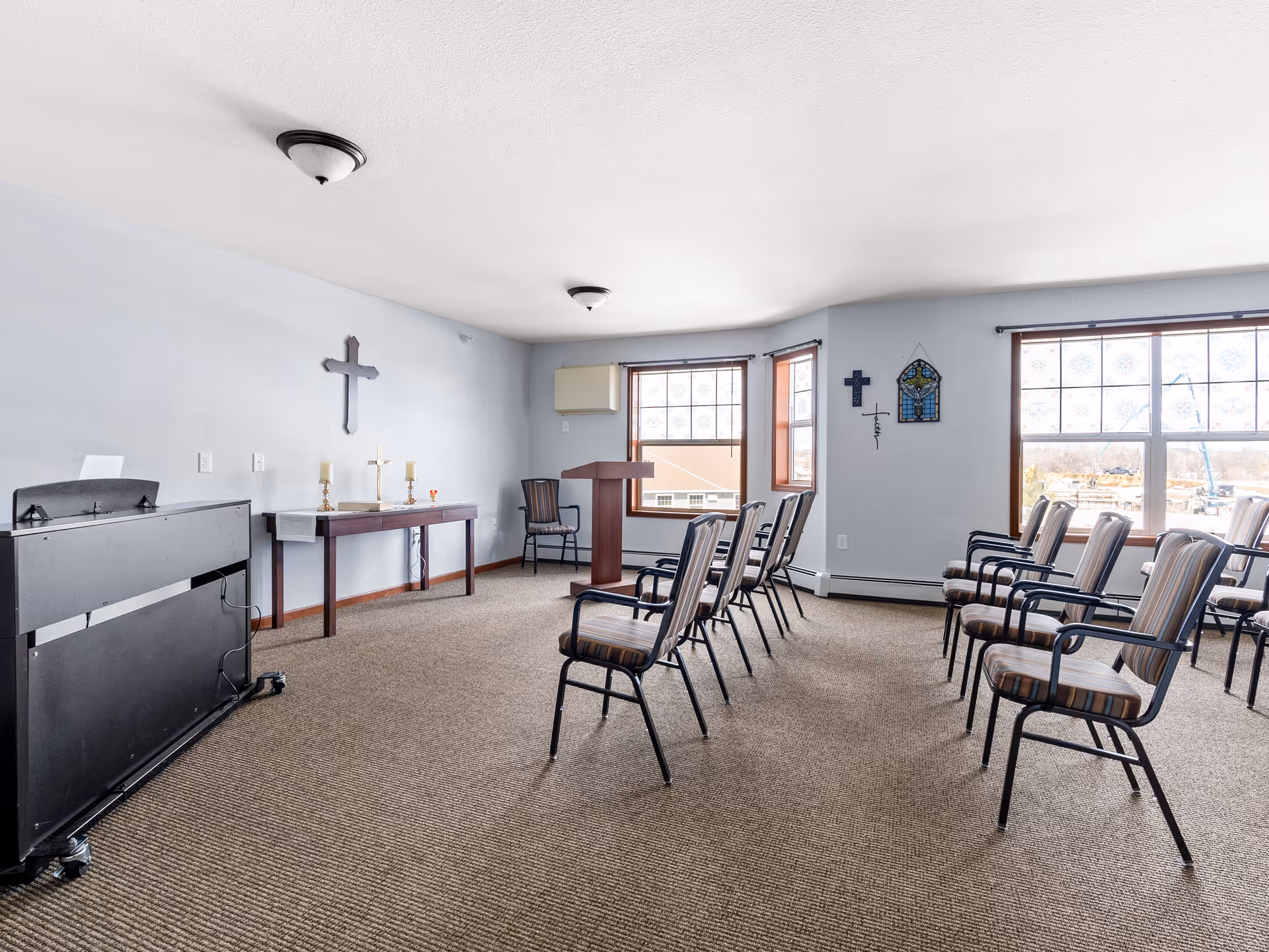 A small chapel or prayer room with rows of chairs facing a wooden podium and a table with religious items including candles and crosses. The room has light-colored walls, carpeted floor, and large windows letting in natural light. Religious crosses and stained glass decorations are mounted on the walls.
