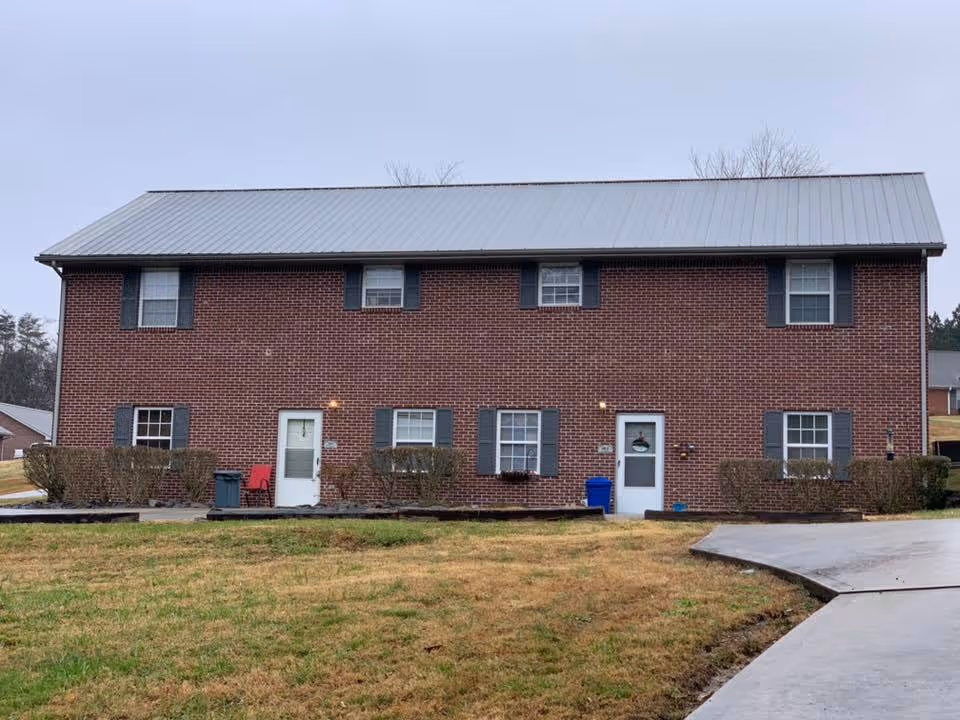 Two-story red brick building with a metal roof, multiple windows and doors, shrubs along the front, and a driveway leading up to it.