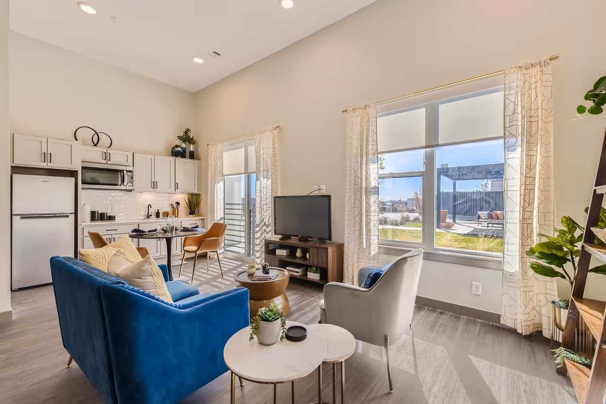 Bright and modern living area in a senior living facility featuring a blue sofa with cushions, a gray armchair, two round white side tables with a small plant, a wooden TV stand with a flat-screen TV, large windows with sheer patterned curtains letting in natural light, and an open kitchen with white cabinets, a refrigerator, microwave, and a small dining table with four chairs.