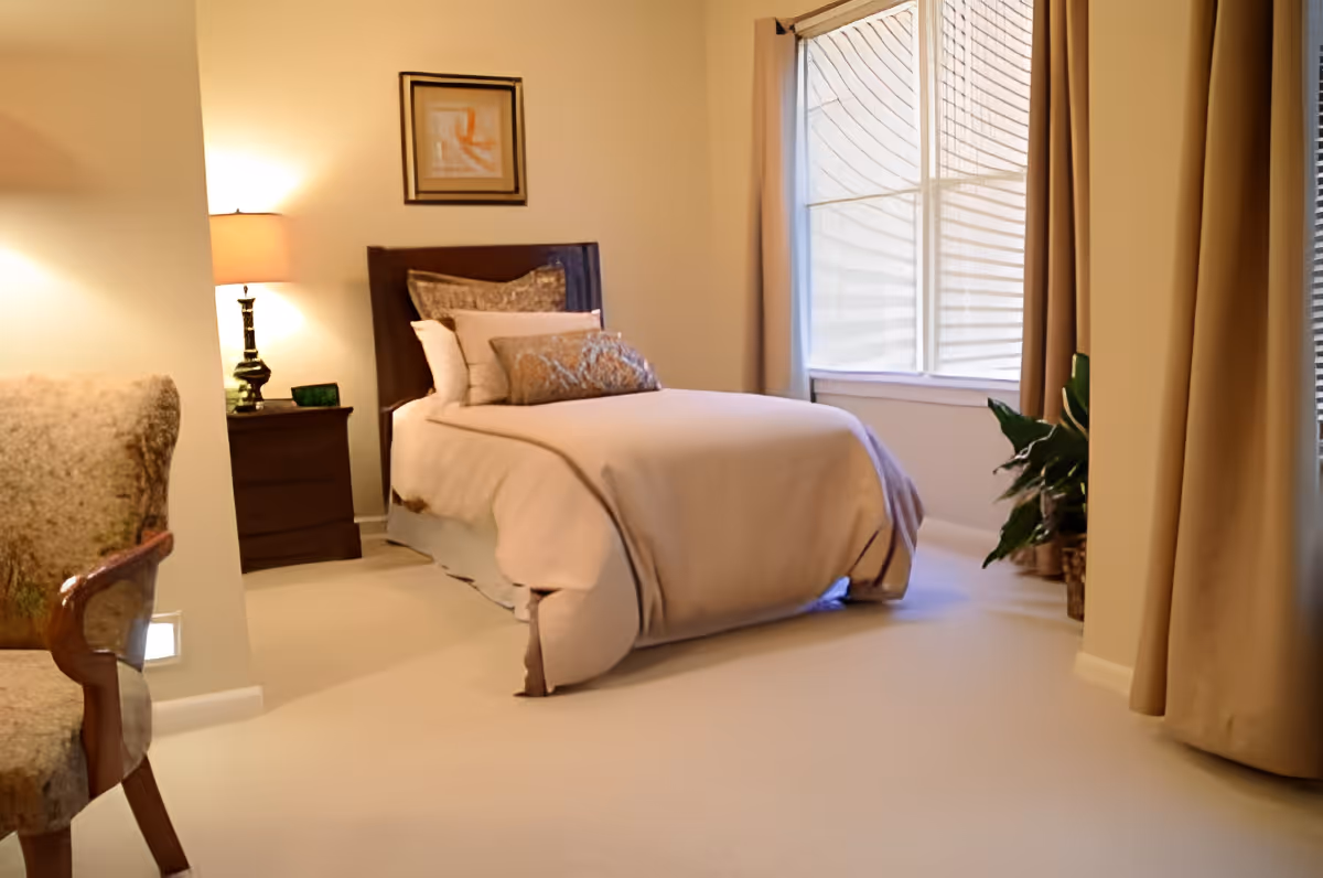 A cozy bedroom with a single bed made up with beige bedding and multiple pillows. There is a wooden nightstand with a lamp and a small decorative item next to the bed. A framed abstract artwork hangs on the wall above the bed. A window with beige curtains lets in natural light, and a green potted plant is placed near the window. A cushioned armchair with wooden armrests is partially visible on the left side of the image.