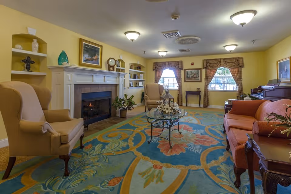 A cozy living room in an assisted living facility with yellow walls, a fireplace, two armchairs, a sofa, a glass coffee table with decorative items, a colorful floral carpet, and windows with plaid curtains letting in natural light.