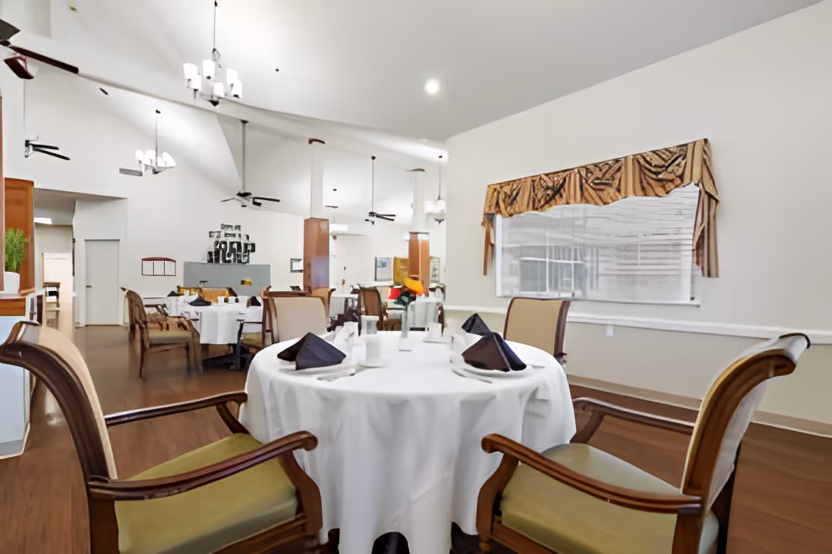 A dining room in Morada Victoria with round tables covered in white tablecloths, set with black folded napkins and white cups. The room features wooden chairs with green cushions, hardwood floors, ceiling fans, and large windows with decorative valances.