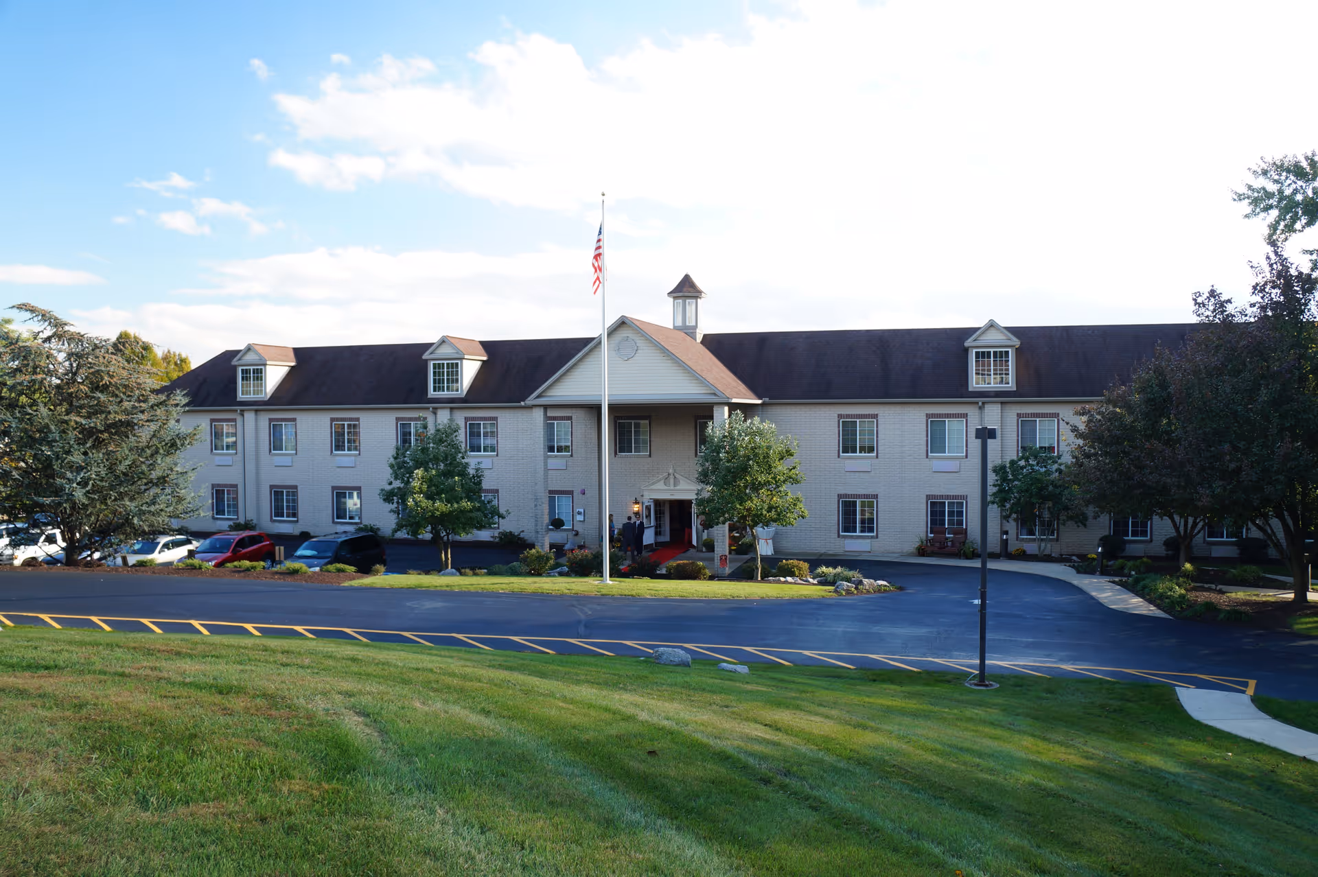Front exterior view of a two-story senior living facility building with a pitched roof, dormer windows, an American flag on a flagpole in front, surrounded by trees and a well-maintained lawn under a partly cloudy sky.