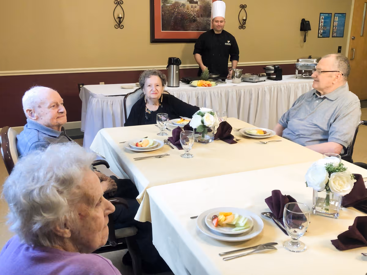 Four elderly individuals seated around a dining table set with plates of fruit and glasses, with a chef standing behind a buffet table in the background inside a dining room.