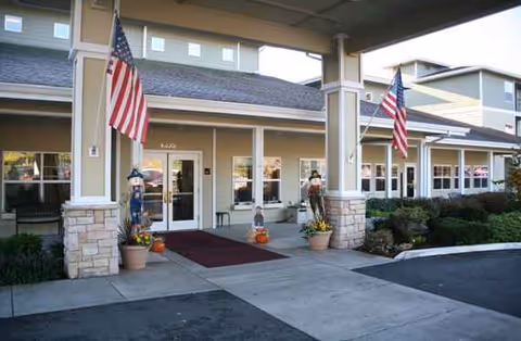 Entrance of a senior living facility with two American flags mounted on pillars. The building has beige siding with stone accents at the base of the pillars. There are seasonal decorations including pumpkins and scarecrows near the entrance doors.